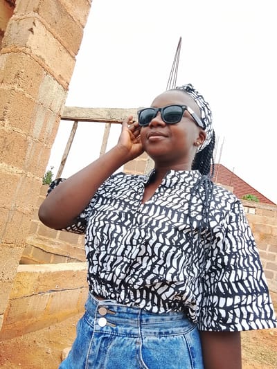 A young woman with dark skin, wearing sunglasses, a black and white patterned top, a blue denim skirt, and a headscarf, stands outside in daylight. Her left hand is raised to her face. The background includes a brick building under construction, steps, and a hint of a red roof on another building. The location is Barikin Sabon Gidan Kanat, Nigeria, based on source information. The image suggests an informal setting.
