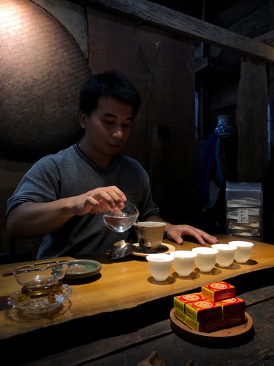 A man, appearing to be of East Asian descent with dark hair and wearing a grey t-shirt, is shown at a wooden table. He is in the process of pouring liquid from a clear glass vessel into a small, ornate tea cup. To his right, there are five small white teacups arranged in a row. In the foreground, on the right side of the table, there are several small, square red and yellow boxes stacked on a wooden tray. On the left side of the table, there is a glass ashtray with a small, green, shallow dish containing loose tea leaves. Behind the man, the wall is textured with woven material. To the far right, a clear plastic bag filled with what appear to be tea packets is visible. The overall setting suggests a tea ceremony or tasting, possibly in B'Lao, Vietnam, given the reference. The lighting is dim, casting shadows and highlighting the objects on the table.