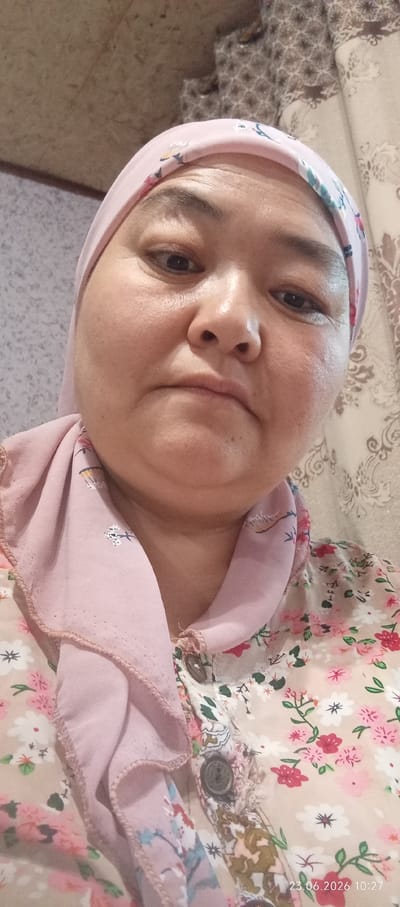 A close-up, low-angle selfie shows a woman with a light pink hijab and a floral-patterned shirt. She is indoors, as indicated by the patterned wallpaper in the background. The lighting is soft and appears to be artificial. The woman's face is the primary focus, with her eyes looking slightly down. The image was taken on June 23, 2026, at 10:27 AM. The location is identified as Chon-Kara, Kyrgyzstan.