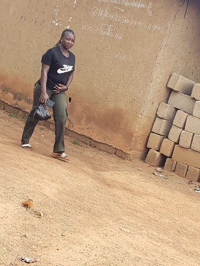 A woman walks on a dirt road in Kachia, Nigeria, in front of a textured, beige wall with chalk markings. She is wearing a black Nike t-shirt and green cargo pants. In her right hand, she holds a black bag. She is also wearing white sandals. To her right, a stack of tan bricks is piled against the wall. The ground is covered in loose dirt and small stones. In the distance, a small bird is visible on the ground. The sky is overcast, suggesting a neutral lighting condition.