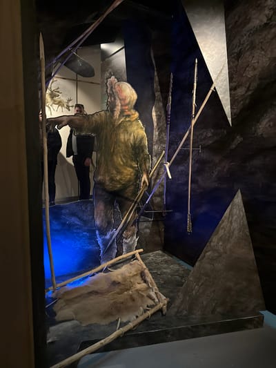 The image displays an indoor museum exhibit located in Vavřinec, Czechia, designed to evoke a prehistoric or primitive setting. A large, two-dimensional cut-out illustration of a male figure, depicted in light brown fur-lined primitive clothing and boots, dominates the central foreground. This figure extends its left arm forward in a pointing gesture and holds several long wooden spears or staffs in its right hand. On the floor in front of the figure, a rudimentary bed-like structure is visible, fashioned from rough wooden poles and covered with a light-colored animal fur hide. The exhibit's backdrop consists of dark, textured panels simulating a cave environment, alongside which two additional long, pointed spears are vertically mounted. A sharp, grey triangular panel forms part of the right side of the exhibit space. A distinct blue light illuminates the lower left area of the scene. In the blurred background, behind the central figure, two dark-clothed live individuals are observing the display, and a panel featuring illustrations of two reindeer or similar antlered animals is visible.