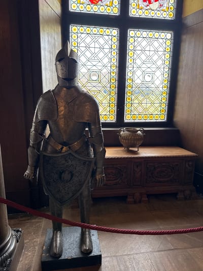 An interior view in Cochem, Germany, features a suit of armor displayed near a stained-glass window. The armor is metallic and heavily ornamented. The stained glass window has a yellow geometric design. A decorative cabinet is visible in the background, along with a brass vase. A thick red rope is draped near the armor. The room has wooden paneling and flooring.