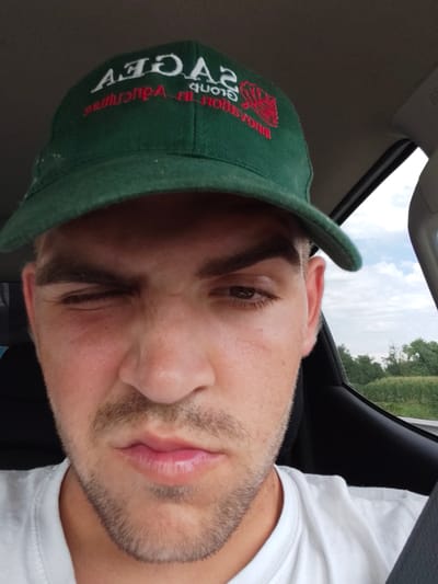 The media file shows a close-up, selfie-style shot of a young man inside a vehicle, likely a car, as suggested by the interior elements like the seatbelt and window frame. He is wearing a green baseball cap with white and red text. The visible text on the cap appears to be a logo with "SAGE" in large white letters and some smaller text in red below.
The man is looking directly at the camera with a somewhat mischievous or playful expression, as indicated by him winking with his left eye and a slight smirk. He has short, dark hair, brown eyes, and a light stubble on his chin and upper lip. He is wearing a white t-shirt. The background visible through the car window suggests an outdoor setting with green trees and a cloudy sky, indicating it's daytime. The overall lighting suggests it could be late morning or afternoon. The scene appears to be a casual moment, possibly while traveling. No specific event is depicted, but the self-portrait suggests the subject is capturing a personal moment.
