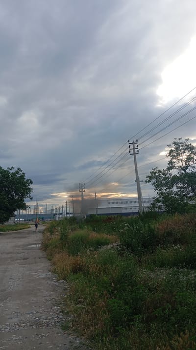 The image depicts an outdoor scene in Baksan, Russia. The setting appears to be an industrial or semi-industrial area with a dirt road in the foreground. Tall grass and various plants grow along the roadside and in the middle ground. In the distance, there is a long, modern building with a white facade and blue trim. Several utility poles with power lines stretch across the frame, indicating infrastructure.

A notable detail is a plume of dark smoke rising from behind one of the utility poles in the middle distance, suggesting a fire or industrial emission. The sky is overcast with dramatic, dark clouds, but there are also hints of bright sunlight breaking through, possibly indicating sunrise or sunset. The overall impression of the weather is cloudy and potentially stormy.

In the foreground, a single person is visible walking on the dirt road, carrying a yellow object, possibly a tool or equipment. The person is small in the frame, emphasizing the scale of the surroundings. The dirt road is uneven and appears to have puddles, suggesting recent rain. The presence of trees on the left and right edges of the frame adds a touch of nature to the otherwise industrial landscape. The time of day is likely early morning or late evening due to the quality of light and the dramatic sky. There is no visible text in the image.