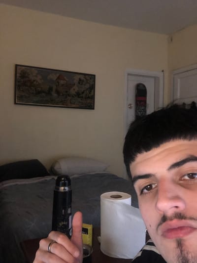The image is a close-up selfie taken in what appears to be a bedroom. The person in the foreground, with dark hair and a mustache, is the primary subject. In the background, there is a bed with a dark gray comforter and pillows. A black thermos is standing upright on a table next to a roll of toilet paper. A framed tapestry depicting a rural scene hangs on the wall above the bed. To the right of the tapestry, there is a doorway with a skateboard leaning against it. The room is illuminated by warm, artificial light, and the walls are painted a light yellow color. There is no visible indication of the city of Beccar, Argentina.