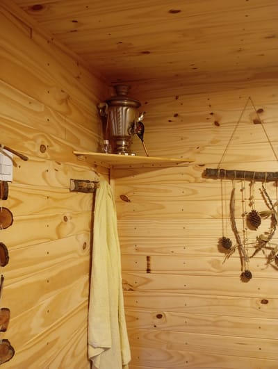 A metal samovar sits on a wooden shelf in the corner of a room with pine-paneled walls and ceiling. A yellow towel hangs from a wooden peg on the wall below the shelf. To the right of the samovar, a string of natural decorations, including pinecones and dried leaves, hangs from a branch. The overall impression is of a rustic or sauna-like environment, possibly in a private residence. The location in Ensenada, Argentina, is not discernible from the image itself.