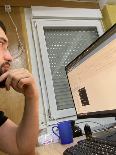 A medium shot captures a man from the chest up, seated at a desk and looking intently at a computer monitor. He is resting his chin on his right hand, his elbow supported by his forearm. His left arm is not visible. The man has dark hair and a beard. The computer screen displays a spreadsheet or database with text in what appears to be Russian. A blue mug, a dark bottle, and a small bottle with a green label are on the desk. A white package with the word "CREATINE" is also visible. The background shows a window with blinds, a wall, and a white electrical outlet with a wire. The scene is indoors, suggesting a home office or workspace. The overall lighting is dim, with a warm tone from an artificial light source.