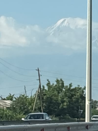 The image depicts a scene captured from a moving vehicle, likely a car, as indicated by the reflection on what appears to be a window. The perspective is from inside looking outwards and upwards. The primary subjects are the sky, trees, a utility pole with wires, and a portion of another vehicle, possibly a white van, visible to the right.

The sky is a pale blue with scattered white clouds, suggesting it's daytime. The clouds are fluffy and somewhat indistinct, with one larger cloud formation on the left having a textured, almost mountain-like appearance.

A dense cluster of green trees occupies the right side of the frame, obscuring much of the background. These trees appear lush and full, indicating a temperate climate during a growing season.

A utility pole stands prominently in the middle ground, with several thin wires extending horizontally across the sky. These wires are characteristic of overhead electrical or telecommunications lines.

To the far right, a section of a white vehicle is visible. We can see part of its side, including a window and a headlight-like illuminated element. This suggests another vehicle is traveling in the same direction or in proximity.

A grey, metallic structure runs vertically along the far right edge of the image, possibly part of the vehicle the photo was taken from, or a roadside barrier. It has three distinct red circular markings.

The image is oriented vertically, with the top of the scene appearing on the left and the bottom on the right. The overall impression is of a roadside view during daylight, possibly in a semi-urban or rural area with natural elements and infrastructure. No people are visible. No text is present in the image. The location context provided is Ayntap, Armenia, which aligns with the presence of trees and utility infrastructure.
