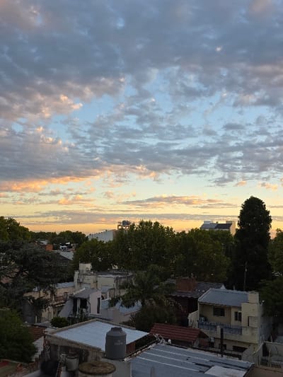An elevated panoramic view depicts a densely populated urban area within Villa San Andrés, Argentina, during a sunset or dusk lighting condition. The sky, occupying the upper two-thirds, features extensive stratocumulus cloud cover. Clouds in the upper sky are predominantly dark grey, interspersed with patches of light blue. Towards the horizon, the clouds are illuminated with warm orange and yellow hues.
The midground and foreground reveal a compact residential landscape. Numerous single-story and low-rise buildings display a mix of architectural styles, characterized by light-colored facades (e.g., white, pale yellow, grey) and various roof types, including corrugated metal, flat concrete, and reddish-brown tiles. Mature trees with dark green foliage are abundant, often rising above the rooftops and forming a prominent green canopy that partially obscures distant structures. A tall, slender, dark green tree stands vertically on the right side of the frame.
On rooftops in the foreground, a prominent grey, cylindrical plastic water tank is visible in the lower-left quadrant. Adjacent rooftops feature other smaller, unidentified cylindrical objects. No individuals, discernible actions, or interactions are present within the scene. The overall environment is static, bathed in the natural light of the setting sun.
