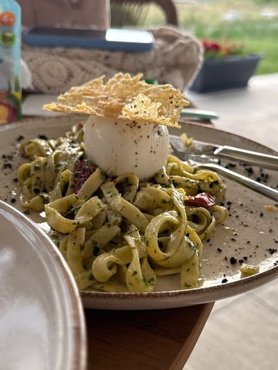 A close-up shot captures a delicious plate of pasta, likely served at a restaurant. The main subject is a generous portion of fettuccine pasta, tossed in a vibrant green sauce, possibly pesto, with visible herbs and what appear to be sun-dried tomatoes. Alongside the pasta is a large ball of burrata cheese, its creamy white exterior contrasting with the pasta's color. A delicate, golden-brown cheese crisp is artfully placed on top of the burrata, adding a textural element. The dish is seasoned with black pepper and some dark specks, which could be truffle shavings or finely chopped olives.
The setting appears to be outdoors or in a room with a view of greenery, suggested by the blurred background of trees and foliage. Sunlight filters in, creating a bright and inviting atmosphere, indicating daytime. The lighting highlights the textures and colors of the food, making it look appetizing.
Other objects visible include part of a white plate with a rustic rim, a wooden serving bowl on the right, and a fork resting on the pasta. A patterned beige fabric or tablecloth is seen on the left side of the frame. In the further background, a dark object, possibly a laptop or tablet, and a blurred glimpse of a chair are discernible.
The overall impression is one of a leisurely meal in a pleasant environment, focusing on the visually appealing presentation of the food. No people are clearly visible. The image evokes a sense of enjoyment and culinary delight. There is no visible text in the image.