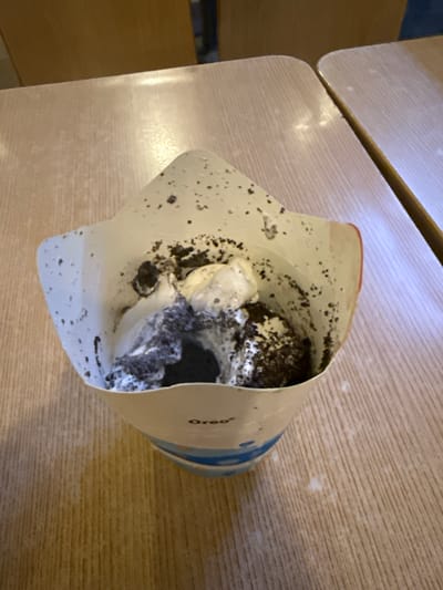The image shows an overhead view of an Oreo ice cream dessert in a white paper cup with a red rim, placed on a light-colored wooden table. The ice cream is mixed with crushed Oreo cookies, and some pieces have fallen onto the inside of the cup. The cup has "Oreo" printed in black on its side. The table has a visible wood grain pattern and appears slightly worn. In the background, out of focus, are parts of wooden chairs and another table. The lighting suggests an indoor setting, possibly a restaurant or cafe. There is no visible indication of the specific location within Manila, Philippines.