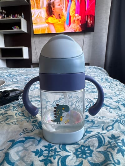 This is a close-up, rotated, horizontal shot of a baby bottle with handles. The bottle is clear with a cartoon dinosaur graphic visible inside. It has a light blue top and a darker blue band around the middle. The handles are a dark blue, curved shape. The bottle is resting on a blue and white patterned fabric, possibly a blanket or tablecloth.
In the background, on the left side of the image, there is a television screen displaying a vibrant image of people in colorful costumes, possibly from a performance. Below the television are shelves containing various items, including what appear to be small bottles or containers. The overall setting appears to be indoors, likely a living room or nursery. There is no discernible text in the image. The lighting suggests it is daytime, but the specific time of day and weather conditions are not apparent. The image focuses on the baby bottle and its immediate surroundings.