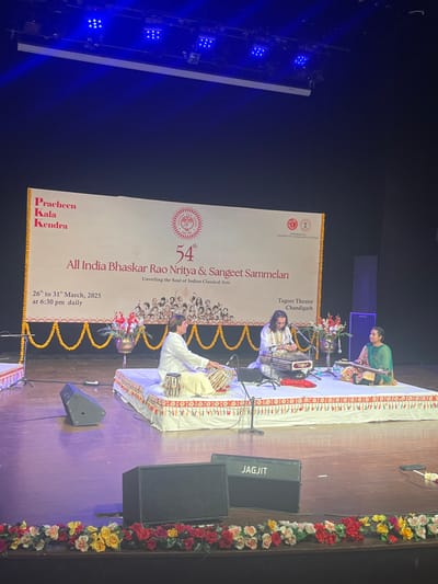 The image shows a stage performance at Tagore Theatre, Chandigarh, India.  The stage is decorated with garlands and flowers, and there is a banner behind the performers that reads, "54th All India Bhaskar Rao Nritya & Sangeet Sammelan Unveiling the Soul of Indian Classical Arts."  There are three performers on stage:  one male seated on the left playing tabla, one male seated in the middle playing a string instrument, and a female seated on the right playing a string instrument.  A large speaker marked "JAGJIT" is on the stage in the foreground.   The performance is part of the 54th All India Bhaskar Rao Nritya & Sangeet Sammelan, which is taking place from March 26th to 31st, 2023, at 6:30 PM daily.  The performance is held at Tagore Theatre, Chandigarh, India, which is located at 30.733 latitude and 76.79 longitude.