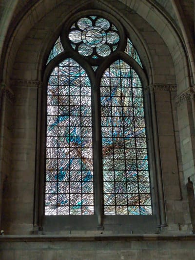 The image shows a tall, arched stained-glass window within a stone structure, likely a church. The window is divided into two large, vertical panes, topped by a circular rose window with multiple small circular panes arranged in a floral pattern. The stained glass depicts abstract patterns of blues, whites, and browns, with a network of dark lead lines dividing the colored glass. The stone architecture surrounding the window is visible, with arched stonework and decorative elements. The overall impression is one of historical grandeur and artistic craftsmanship. The image does not contain any people or identifiable subjects. This stained-glass window is located in the Church of Saint-Nicolas in Urvillers, France.