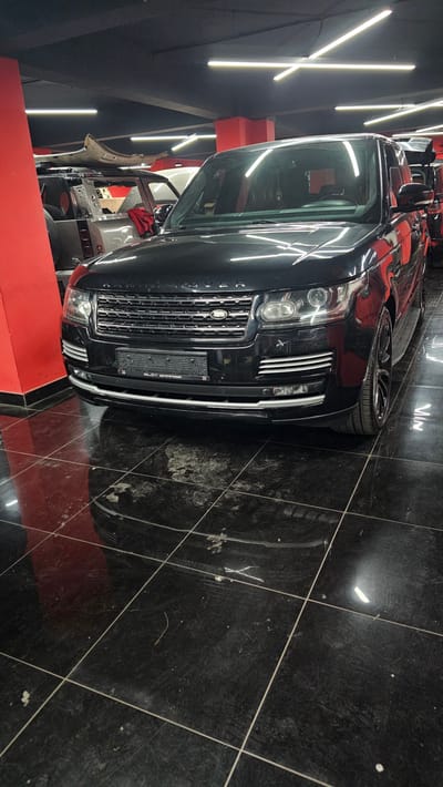A black Range Rover is parked in a garage with a red pillar on the left. The garage has a dark ceiling with modern LED lighting and a black tiled floor. Other vehicles are visible in the background, one of which appears to be undergoing some kind of work, with its hood open and parts exposed. The Range Rover has its headlights on and the front grille is prominent. The license plate is visible. The overall impression is of an indoor automotive service or display area. The location is Ataşehir, Turkey.