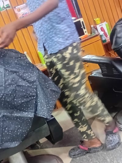 A blurred indoor image depicts a barber attending to a client in what appears to be a barbershop in the city of Dorayi, Nigeria. A standing individual, partially obscured by motion blur, wears a light blue patterned shirt and camouflage trousers with dark grey/black Crocs featuring pink straps. Their right arm is raised, extending toward the seated client. The client is covered by a black cape with small white polka dots. In the background, a wooden paneled wall supports posters, one depicting a green scooter and another a red car. A wooden counter holds various items, including a white appliance, green bottles, and a red "V-MARK" grooming tool. A black barber's chair is visible on the right. The floor features light and dark patterned tiles.