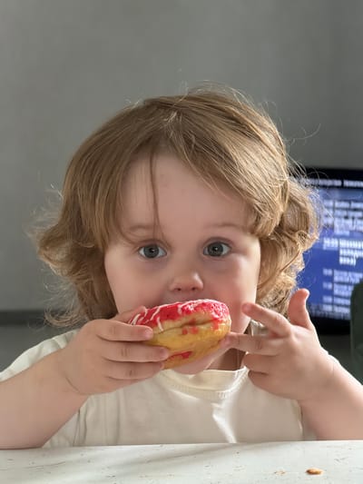 A young child with light brown hair is holding a donut with red icing and white stripes and bringing it to their mouth. The child is wearing a white t-shirt. The background shows a wall and a blurred television screen displaying text. The scene appears to be indoors, likely in a home setting, possibly in the city of Kamyennoye, Russia. There is a white table in front of the child.