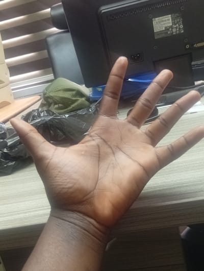 A dark-skinned hand with its palm facing the camera is held up in the foreground. The hand is open, with fingers spread. Behind the hand, a black plastic bag is visible, and further back, a green mesh cap lies on top of it. A portion of a computer monitor and blinds are visible in the background, suggesting an indoor setting. The scene is likely within a home or office environment in Shiroro, Nigeria, as per the prompt.
