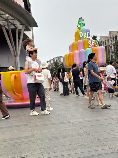 In the Haidian District of China, a father carries his daughter on his shoulders while walking on a paved plaza. The girl, wearing a light pink t-shirt, looks to her left. The father, dressed in a white t-shirt and dark pants, holds a white and red paper bag in his right hand. Behind them, a large, colorful sculpture resembling a teacup is visible. Further in the background, a multi-tiered, brightly colored structure displays the words "DREAM SEEK" in neon letters. A crowd of people strolls through the plaza, some carrying bags and others engaging in conversation. The environment suggests an outdoor shopping or entertainment area, with modern buildings visible in the distance under an overcast sky.