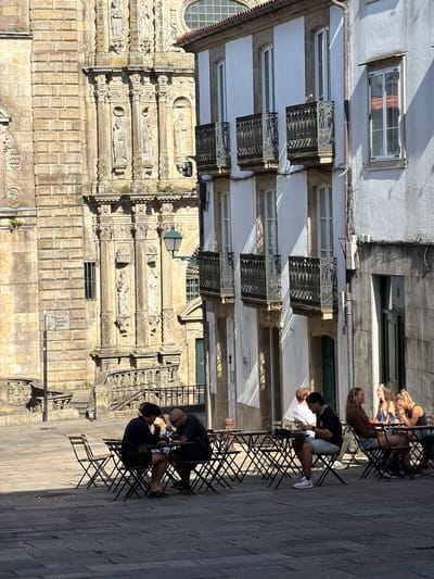 The image depicts an outdoor cafe scene on a sunlit day in Santiago de Compostela, Spain, likely near the Cathedral of Santiago de Compostela. The foreground shows several dark metal tables and chairs set up on a cobblestone plaza. Three groups of people are seated at these tables. On the left, a couple is engaged in conversation, both looking down at their table. In the middle, a man in a black and white striped shirt and light shorts is looking at his phone. To the right, three women are seated, with two appearing to be in conversation and the third looking away.
Behind the cafe, an ornate, aged stone building with intricate carvings and statues dominates the left side of the frame. This building, with its arched doorways and textured stonework, suggests historical architecture. To the right of this, a white building with multiple windows and decorative black metal balconies stands out. The architecture suggests a European city with a rich history. Shadows cast by the buildings and the tables indicate the time of day is likely midday or early afternoon. A street lamp is visible on the stone building, and a street sign is mounted on a pole.