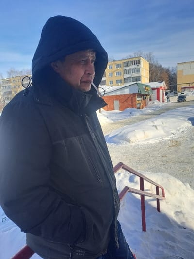 This image depicts an outdoor winter scene in Bolshaya Elhovka, Russia. In the foreground, a male individual, appearing middle-aged, stands with a dark hooded winter jacket drawn up over his head. He has his hands in his jacket pockets and a lit cigarette in his mouth, oriented towards the right side of the frame. Below and to his right, red-painted metal railings are partially obscured by snow. The ground is extensively covered in white snow. In the mid-ground and background, under a clear blue sky, multiple structures are visible: a light yellow multi-story apartment building, a smaller commercial building with a red brick base and a green roof, and another lighter-colored building to the far right. A snow-covered paved area, likely a road or parking lot, extends into the distance on the right, where a dark vehicle and a pedestrian are faintly discernible. Bare trees are visible on a distant hill or embankment beyond the buildings.