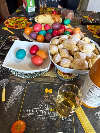 A high-angle shot captures a table laden with food and beverages, suggesting a festive or celebratory gathering. A large white bowl overflowing with powdered pastries dominates the upper center. Nearby, a glass filled with a light-colored liquid and a bottle, partially visible, add to the impression of refreshments. A smaller white bowl containing a few dyed Easter eggs is situated near the larger bowl of pastries. To the left, a basket holds an assortment of colorful Easter eggs, accompanied by sliced bread and a wrapped loaf.
The table is covered with a patterned tablecloth, featuring images of pizza, pasta, and culinary-related text. A pizza cutter with a wooden handle rests on the tablecloth. The background reveals a person partially visible on the left side of the frame.
The text on the beverage bottle is partially visible, with phrases like "Energiina" and "coca-cola.bg" suggesting it's a commercial product. Text in Bulgarian, including the address of "ул. Кана Халония Ба", indicates the location is likely in Bulgaria. Given that Rosen is a common name in Bulgaria, without additional context, it cannot be definitively determined that this image was taken in the city of Rosen, Bulgaria.