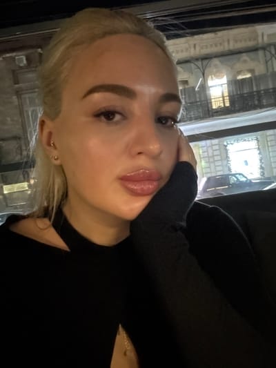 A woman with fair skin and blonde hair is posing for a photo. She has full lips and defined eyebrows, wearing a black top and a delicate gold necklace. Her hand is near her face. The background reveals an urban setting at night with buildings visible through a window. The overall lighting creates a soft, flattering glow on her face.