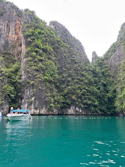 A boat with people on it is anchored in a bay with steep, tree-covered cliffs on either side. The water is a vibrant turquoise color. The boat has the name "SAWANDA HAEL" on its side. There are several buoys in the water, indicating a swimming area. The sky is overcast. The scene appears to be a tropical bay, likely in Southeast Asia.