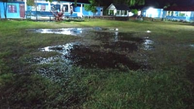The image depicts an outdoor scene at night, likely in Uteunreutoh, Indonesia. A grassy field with several large puddles of water occupies the foreground and midground. The water reflects the light from nearby buildings, creating a shimmering effect on the surface. In the background, a row of blue and white buildings with white trim are visible. Some of these buildings appear to be multi-story. One building on the far right has a dark, gabled roof. There is also a small, white structure with a dark roof in the midground, resembling a gazebo or small shelter. The lighting suggests artificial illumination from the buildings, casting shadows and highlighting the wet areas of the field. Vegetation, including trees and bushes, is present around the buildings and in the background. There are no people or animals clearly visible in the image. The overall impression is of a wet, outdoor area adjacent to a complex of buildings, possibly a school or community center, at night.