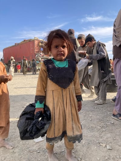 The image captures an outdoor scene in Chaman City Tehsil, Pakistan, set on an unpaved, dusty, and gravelly surface under a clear blue sky with minimal cloud cover.
In the foreground, a young child, approximately 3-5 years old, stands centrally, facing forward. The child has dark, shoulder-length hair and is barefoot, with visible dust on the lower legs and feet. Clothing consists of a light tan or brown long-sleeved dress featuring black, possibly beaded or embroidered, trim around the squared neckline, cuffs, and in vertical lines down the front, worn over a teal undershirt. The child holds a black plastic bag in the left hand.
To the immediate left of the central child, the lower arm and bare foot of another individual, dressed in light brown attire, are partially visible. Further back on the left side, a red semi-trailer truck is parked, heavily loaded with various items, some covered by netting. Several adult males are present in the mid-ground near the truck; one wears a white turban and light-colored clothing, while others are in darker traditional garments. A smaller child in a red top is also discernible among these individuals.
In the mid-ground to the right of the central child, two boys are observed. One boy, wearing a light grey traditional outfit and a light-colored cap, holds a white bowl. The second boy, dressed in a darker grey garment and a dark knitted cap, is handling a clear plastic bag, seemingly engaged in distributing or receiving items. On the far right, the lower leg and foot of another person, wearing a purple garment and dark sandals, are partially visible.