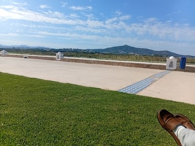 A wide-angle, eye-level shot captures a serene outdoor scene in Martil, Morocco, under a partly cloudy blue sky. The perspective is from a grassy area where someone reclines, their feet adorned with brown leather loafers and white socks visible in the lower right corner.
The immediate foreground is vibrant green grass, smoothly transitioning to a concrete pathway. This pathway is punctuated by decorative, white Moorish-style posts. Beyond the pathway is a low stone wall that separates the path from a stretch of land.
In the distance, a blend of urban development and natural terrain unfolds, including buildings in the city, a range of hills, and a prominent mountain in the background. The skyline is accented by the scattered clouds.
The overall mood of the image is tranquil, suggesting a moment of relaxation in a scenic locale.