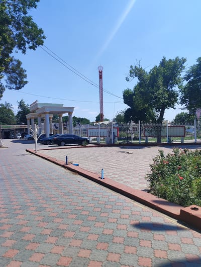 The image shows a wide, paved courtyard in Tursunzoda, Tajikistan, under a clear blue sky. On the left, a large white building with a colonnade entrance is visible, with cars parked in front of it and trees lining the street. A tall, red lattice tower stands in the background behind a decorative white fence. A section of green bushes and flowers is in the lower right corner. The courtyard is paved with interlocking grey and red hexagonal tiles.