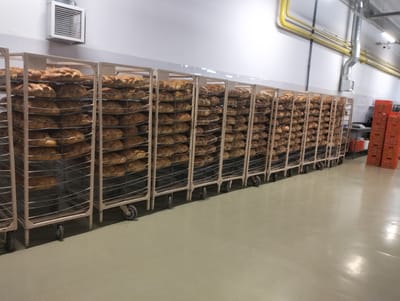 The image depicts a bakery or food production facility in Șoldănești, Moldova. The primary focus is on rows of industrial racks laden with freshly baked bread, arranged on multiple shelves. These racks, constructed with metal frames and wire shelves, are positioned on wheels, suggesting mobility within the facility. The bread appears to be of a light brown, uniform color and shape, possibly loaves or rolls. In the background, a white wall with some ventilation systems and yellow pipes are visible, indicating an industrial setting. To the right of the racks, several stacked orange plastic crates are present. The floor is a smooth, light-colored surface, reflecting light in some areas. No people are visible in the frame. The overall environment appears clean and organized, consistent with a commercial food preparation area.