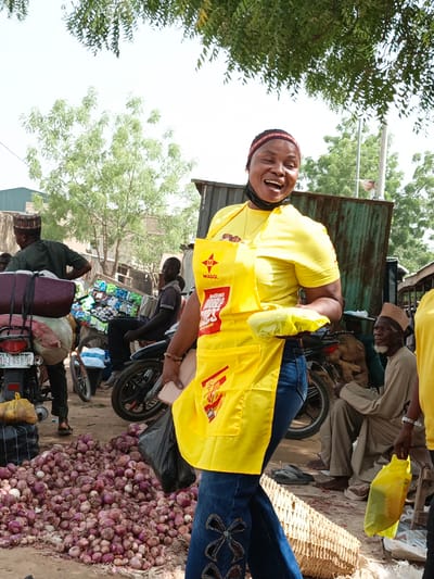 This vibrant image captures a bustling outdoor market scene in Kawaji, Nigeria, under bright, clear daylight. The central figure is a cheerful woman, smiling broadly with an open mouth, exuding enthusiasm. She wears a bright yellow t-shirt, blue jeans with distinctive embroidered patterns, and a prominent yellow apron. The apron clearly displays the "MAGGI" logo and "DOUBLE STAR" branding, suggesting she is engaged in a brand promotion or sales activity. A black face mask is pulled down to her chin, and a colorful patterned headband adorns her head. She holds a yellow plastic bag in one hand and a phone in the other.
The market environment is characterized by active commerce. In the immediate foreground, a large, sprawling pile of red onions dominates the ground, indicating the sale of fresh produce. Surrounding the woman are various market-goers and vendors. To her left, a man in a dark green shirt stands beside a motorcycle heavily laden with large sacks and luggage. Another man is seated on a separate motorcycle, which is piled high with numerous small, brightly packaged items. On the right, an older man with a white beard and a traditional cap sits, observing the lively scene. Partially visible on the far right is another individual, also dressed in a yellow top, possibly part of the same promotional team.
The background reveals typical market infrastructure, including simple structures made of corrugated metal and verdant trees under a bright sky. A visible motorcycle license plate, "KANO JJJ 5500", firmly places the scene within the Kano State of Nigeria, consistent with the Kawaji location context. The overall impression is one of a busy, community-focused market bustling with daily life and commercial activity.