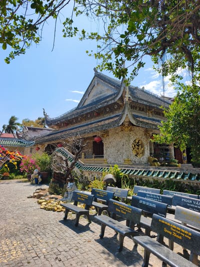 The image depicts a serene courtyard scene in front of a Buddhist temple in Tuy An Nam, Vietnam. The temple, constructed with light-colored stone and adorned with intricate decorative elements, features a multi-tiered, ornate roof with dark tiles. Golden dragon motifs are visible on the roofline. The facade of the temple is decorated with a circular golden emblem. In the foreground, a row of stone benches, inscribed with what appear to be names and dates in Vietnamese, are arranged on a paved courtyard. A statue of a tiger with its cub is positioned near the benches. Lush green foliage and flowering plants surround the temple and courtyard. The sky above is a clear blue with scattered white clouds, and branches of a large tree extend into the frame from the top, casting shadows. A woman in a yellow top and dark pants is visible standing near an entrance to the temple.