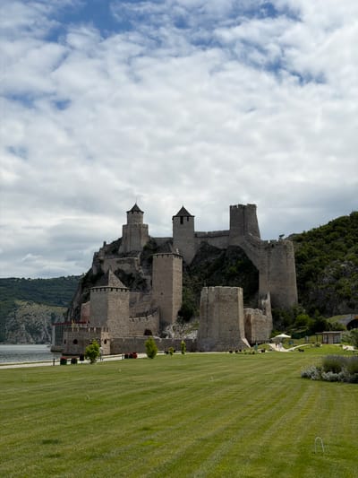 This is a wide-angle, exterior, eye-level shot of Golubac Fortress, located in Golubac Municipality, Serbia. The fortress, constructed of stone, is perched on a rocky cliff overlooking a body of water, likely the Danube River. The fortress features multiple towers, walls, and battlements. In the foreground, a neatly manicured green lawn is visible, with several small white U-shaped wickets scattered across it, suggesting a croquet game in progress or recently played. A few people are walking on a path near the fortress entrance, and some small trees and flower beds are present along the edge of the lawn. The sky is partly cloudy with blue patches. The overall scene depicts a historical landmark situated in a natural, scenic environment, with a recreational area in the foreground.
