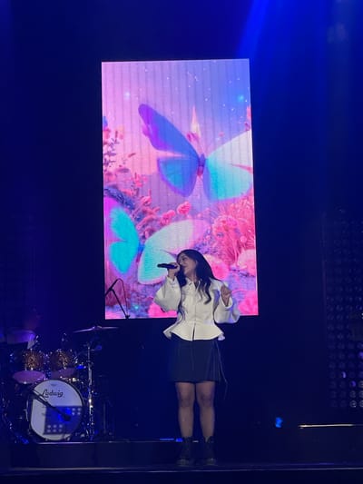 A woman in a white, long-sleeved shirt and black skirt stands on a stage, singing into a microphone. Behind her is a large screen displaying a vibrant image of butterflies and flowers. To the right, a Ludwig drum set is visible. The location appears to be a concert setting, possibly in Salembaran, Indonesia.
