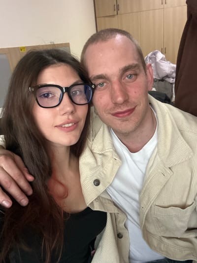 Two individuals, one female and one male, are depicted in a close-up indoor photograph taken in Huangdao District, China. The female is positioned on the left, with long dark brown hair and dark-framed eyeglasses. She is wearing a black top. The male, on the right, has short light brown hair and light blue eyes, and is wearing a white t-shirt beneath a light beige corduroy jacket. The male's left arm is around the female, with his hand resting on her left shoulder. Both individuals are looking towards the camera with slight smiles. The background includes a light-colored wall on the left, a light-colored doorframe with a yellow sticker in the upper left, and a stack of light-colored wooden cabinets in the upper right. A pile of light-colored fabric or clothing is visible below the cabinets on the right, next to a darker, possibly brown, hanging garment.