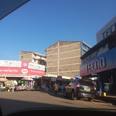 The image captures a vibrant street scene on a bright, sunny day, likely in a commercial area of Sheywe ward, Kenya. The clear blue sky and strong sunlight suggest morning or early afternoon.
In the foreground and mid-ground, the street is bustling with daily activity. On the left side, an array of market stalls operates under colorful awnings, displaying various goods for sale. Several people, including vendors and shoppers, are visible near these stalls. Above the market area, prominent red and white signs read "Happy better life itel" and "itel," with "MACCH COMMUNICATION" and a partial "SAMSUNG" sign further along. Below, a small sign reads "BOUTIQUE."
On the right side of the street, large blue and white commercial signs dominate, featuring "CITY WAY," "TECNO" (twice), and "Infinix," indicating electronics or mobile phone stores. A dark-colored SUV, identifiable by its Kenyan license plate "KDB 695M," is parked curbside, facing away from the camera. A pedestrian in dark clothing walks on the pavement near the vehicle.
In the background, several multi-story buildings with concrete facades rise above the street, some appearing weathered, while others show more modern elements like blue-tinted windows. The overall scene depicts a busy urban or semi-urban commercial hub, characterized by active commerce and everyday street life. The photograph is taken from inside a vehicle, with a slight glare visible in the top left corner, indicating the viewer's perspective.