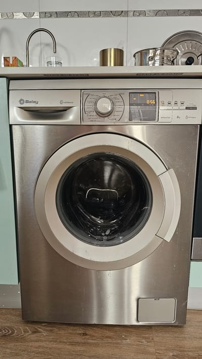 This is a medium-shot, eye-level, exterior, frontal view of a Balay washing machine. The machine is stainless steel and is positioned on a wooden floor against a tiled wall and countertop. The door of the washing machine is closed, and the drum is visible through the glass. The control panel of the washing machine is at the top, featuring a dial for selecting wash cycles, a digital display showing "0:58", and various buttons. To the left of the washing machine, a faucet extends from the tiled wall over a glass containing water and two small cartons. A brass-colored cylindrical object and a metal pot with a lid are placed on the countertop. The overall environment appears to be a domestic setting, possibly a laundry room or kitchen, in María De Huerva, Spain.