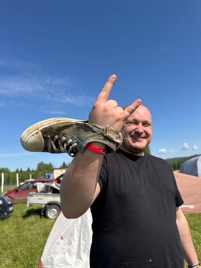 The image displays an outdoor scene in Большая Кивара, Russia, under a clear blue sky with minimal cirrus clouds. A light-skinned male with a bald head and a beard is positioned in the foreground, smiling towards the viewer. He wears a black t-shirt and a red wristband on his right wrist. His right arm is raised, forming a 'devil horns' hand gesture, with a heavily soiled, light-colored canvas sneaker suspended horizontally on his forearm, just above the wristband. A small yellow mark is present on the shoe's toe cap. The background features a grassy field containing multiple vehicles, including a partially visible red car, a small utility trailer with a white cover, and a dark-colored car. Several tents are also present, with a white tent partially visible in the lower-left foreground and a larger brown tent in the mid-ground on the right. Distant green trees and rolling hills define the horizon.