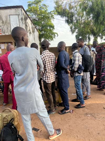 An outdoor scene in Gbaza, Nigeria, depicts a gathering of approximately twelve to fifteen individuals, predominantly adult males of African descent, with at least one adult female visible. Most individuals are standing, appearing to form a loose queue or congregation.

In the foreground, an adult male, viewed from the back, wears a light grey long-sleeved tunic and matching trousers, with his arms crossed. He wears light-colored sandals. To his immediate left, another adult male in a crimson long-sleeved shirt is partially visible, standing near a dark wooden table. To the right of the central foreground figure, an adult male wears a brown and white checkered long-sleeved shirt, dark trousers, and a dark baseball cap.

Further right, a line of individuals extends, including several males and one female wearing a floral hijab and long dress. Many individuals wear sandals or flip-flops. Two men in the mid-ground carry backpacks: one black backpack on a man wearing a grey tunic and light grey trousers, and a tan/khaki backpack on a man wearing a plaid shirt and distressed blue jeans. Another man wears a dark blue long-sleeved tunic and dark trousers.

To the left, a single-story white building with a brown roof exhibits weathering, discoloration, and peeling paint. A white banner or poster, partially inverted, is affixed to the wall, displaying words such as "PRODUCTS," "SUGAR," and "CORN." Below it, a bulletin board with various pinned papers is visible.

The ground is unpaved, consisting of light brown earth or sand, with scattered debris including small pieces of paper and dried leaves. Lush green trees are visible in the background against a bright, slightly cloudy sky, indicating daytime conditions. A low, weathered concrete wall runs across the middle ground, separating some individuals from the immediate foreground. The overall scene suggests an informal outdoor gathering or waiting area.