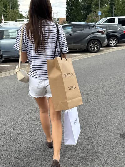 A woman in a striped shirt and denim shorts is walking on an asphalt surface. She's carrying shopping bags, one with "BOTTEGA VENETA" printed vertically and a white "PRADA" bag. The woman has long, dark hair and is wearing dark shoes. Several cars are parked along the side. The environment appears to be an outdoor parking area.
