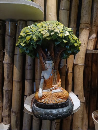 A statue of Buddha is seated under a tree with green leaves. The statue is adorned in orange robes with intricate patterns and golden embellishments. The Buddha is in a meditative pose, with eyes closed and hands resting on its lap. The statue is placed on a lotus-shaped base, which is on a circular platform. Behind the statue, there is a backdrop of vertically arranged bamboo poles. The overall scene suggests a serene and spiritual ambiance. The specific location in Kurnool, India, is not discernible from the image.