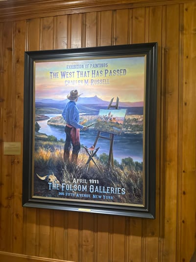 The framed print advertises an "Exhibition of Paintings" titled "The West That Has Passed" by Charles M. Russell. An artist in cowboy attire stands by an easel, painting a scene of cowboys on horseback. The setting appears to be a river landscape with mountains in the background. The text indicates the exhibition occurred in April 1911 at The Folsom Galleries, located at 396 Fifth Avenue in New York. A cow skull is visible in the foreground, adding to the Western theme.