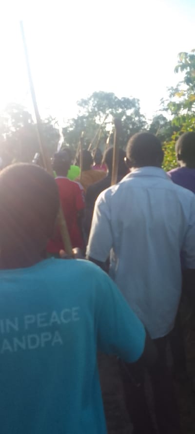 The image shows a group of people in a procession, with the individual in the foreground wearing a teal blue shirt that reads "IN PEACE ANDPA". The individuals in the background are carrying sticks and appear to be walking in a line. The scene is in a rural setting with trees in the background. The sky is bright and there is a lot of sunlight. The location is near Isukha ICHINA, Kenya, based on the provided coordinates. The individuals in the background are mostly obscured and out of focus. The overall feel of the image is peaceful and somber.
