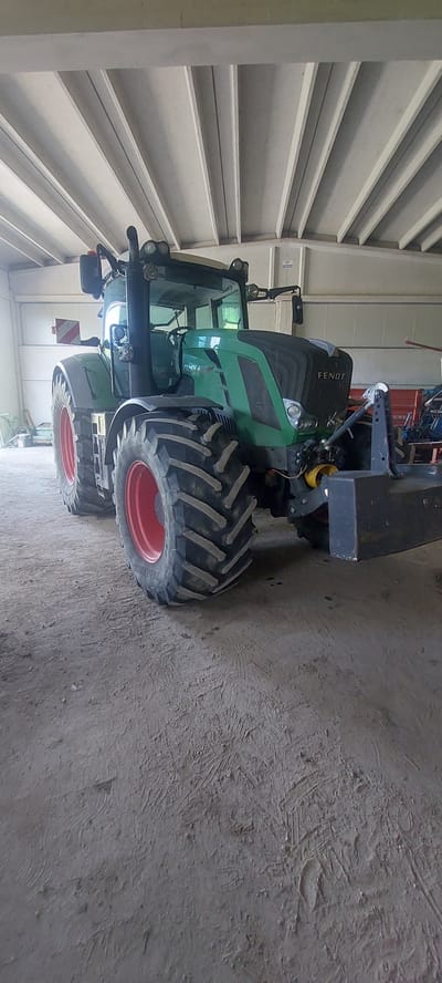 A green Fendt 828 Vario tractor is parked inside a covered structure. The tractor has large, rugged tires with red rims. It is oriented with its front facing slightly to the right of the camera, and its left side is more visible. A black counterweight is attached to the rear of the tractor. The interior of the structure appears to be a barn or workshop, with a concrete floor and a corrugated roof supported by beams. In the background, other farm equipment and supplies are visible, including what appears to be red machinery and possibly a pallet. The lighting suggests it is daytime. The location is identified as Pontestura, Italy.