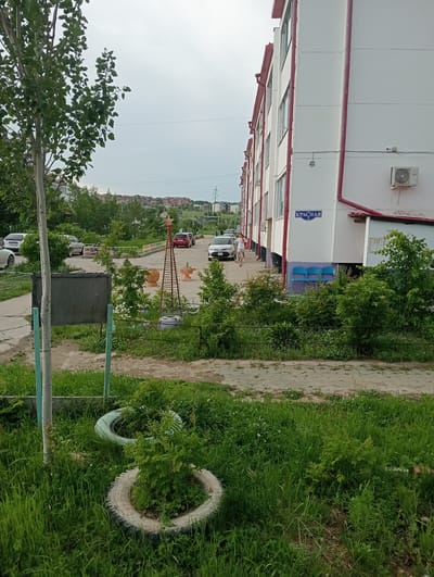 The image depicts a residential area in Chigiri, Russia, on an overcast day. In the foreground, tall green grass is present, with two car tires serving as planters for small bushes. A grey metal board on a light blue metal post stands to the left. A sidewalk paved with light grey concrete slabs runs through the scene. A white multi-story apartment building with red trim is prominently featured on the right side of the image. A street sign reading "КРАСНАЯ 3" (Krasnaya 3) is affixed to the building's facade. Several blue plastic chairs are visible near the building's entrance. In the middle ground, parked cars, including a white SUV and a red sedan, are visible on a road. Further in the background, a line of residential buildings and trees extends towards a distant hill. Two individuals, one wearing a white top and shorts and the other in a white shirt and shorts, are walking on the sidewalk near the apartment building.