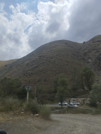 The image depicts a mountainous landscape with a dirt road in the foreground. Several cars are parked on a paved area to the right, adjacent to a weathered structure. The mountainside is covered in sparse vegetation and rocks. The sky is filled with dramatic clouds. A traffic sign is visible on the left side of the road, and the area around it is littered with trash. The scene appears to be a roadside stop or parking area in a rural, mountainous region, possibly in or near Karangisay, Uzbekistan. There are no people visible in the image.