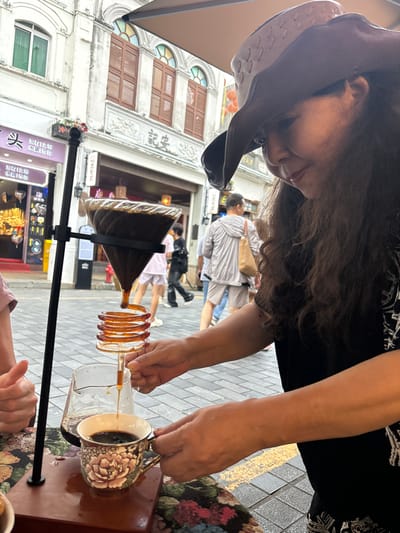 An outdoor scene on a street in Meilan District, China, shows a woman wearing a brown cowboy hat and dark clothing, preparing a pour-over coffee. She is in the foreground, with her hands carefully manipulating the coffee dripper and cup. The coffee is dripping from a conical glass dripper into a decorative ceramic mug with a floral pattern. A glass carafe with brewed coffee sits nearby. In the background, a street lined with traditional architecture is visible. People are walking on the sidewalk, and shop signs with Chinese characters are displayed on the buildings. The ground is paved with interlocking bricks, and a yellow stripe marks a pedestrian pathway. The lighting suggests it is daytime.